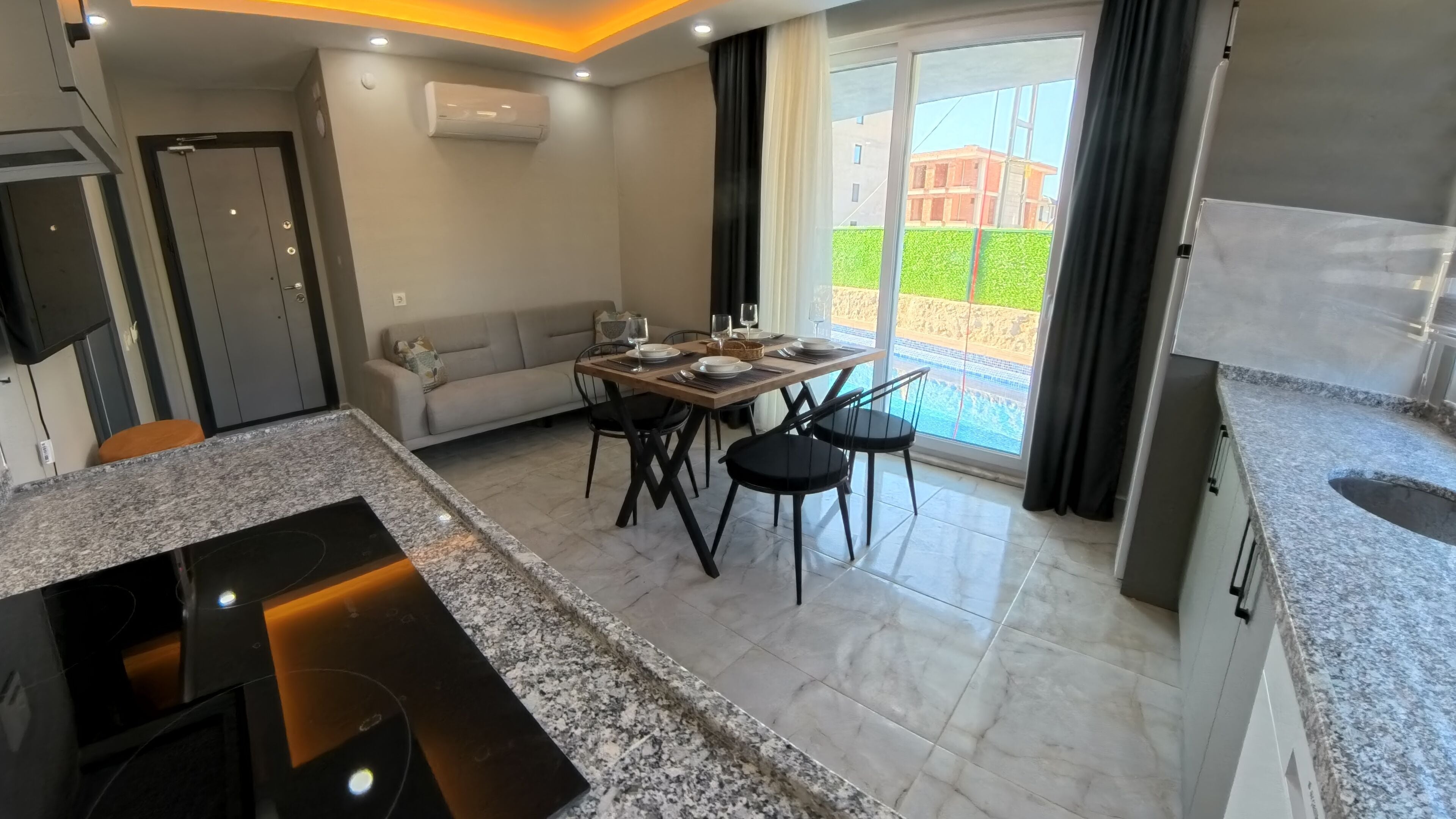 A modern kitchen with granite countertops, a dining area with a table set for four, and a large sliding glass door leading to a balcony with a view of a building and greenery.