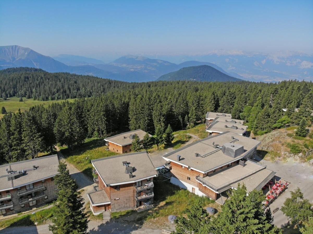 A cluster of modern buildings with flat roofs, surrounded by dense forest and mountains in the background.