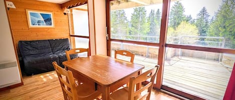 A wooden dining table with four chairs, a black sofa, and a large window with a view of a deck and trees.