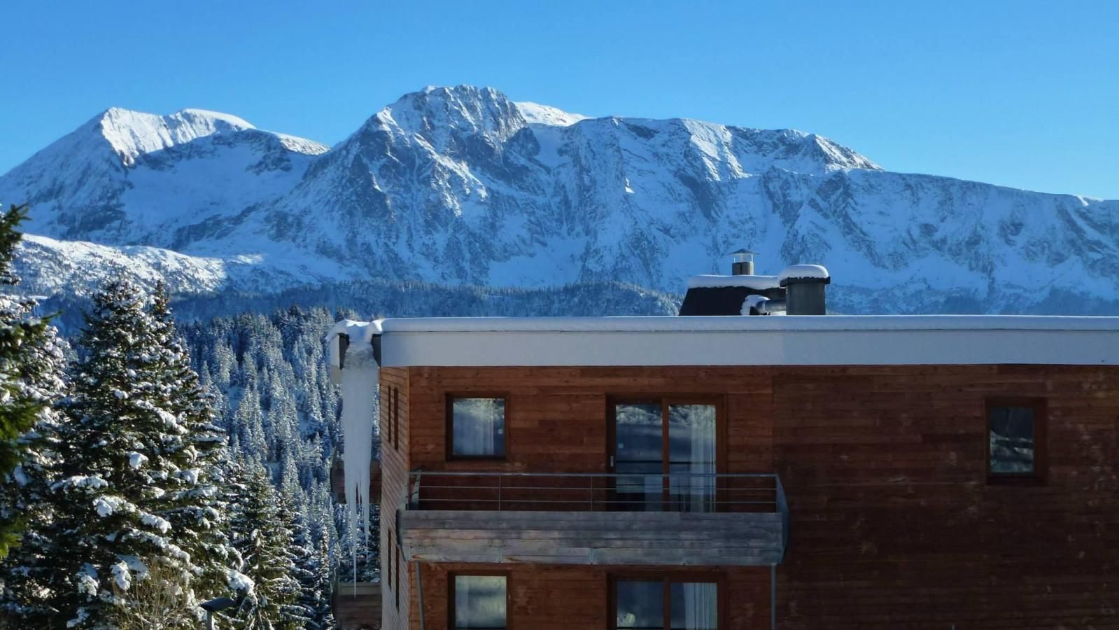 A modern building with a wooden facade and a balcony, set against a backdrop of snow-covered mountains.