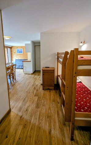 A room with a bunk bed, a small wooden cabinet, and a dining area with chairs and a table.