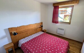 Un dormitorio con cabecera de madera, una cama individual con colcha floral roja, una mesita de noche pequeña, un radiador de pared y una cortina roja en la ventana.