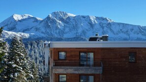 Un edificio moderno con fachada de madera y un balcón, frente a un paisaje de montañas nevadas.