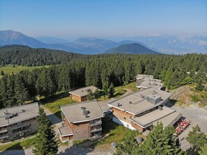 A cluster of modern buildings with flat roofs, surrounded by dense forest and mountains in the background.