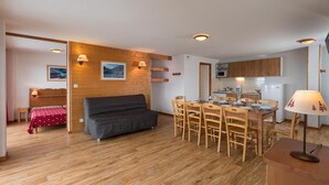 Um quarto acolhedor com piso de madeira, um sofá, uma mesa de jantar com cadeiras, uma cozinha compacta com micro-ondas e uma cama com colcha vermelha.