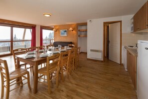 A dining area with a wooden table set for six, wooden chairs, a red curtain, and a view through large windows.