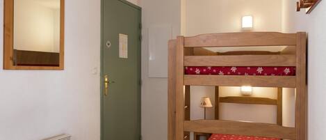 A bunk bed with a red patterned bedspread, a green door with a brass handle, a wall-mounted mirror, and a radiator.