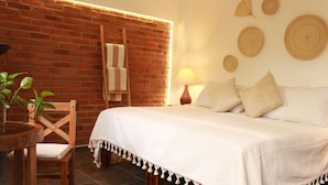 Une pièce avec un mur en briques, un lit avec une literie blanche, une chaise en bois, une petite table avec une plante et une lampe.