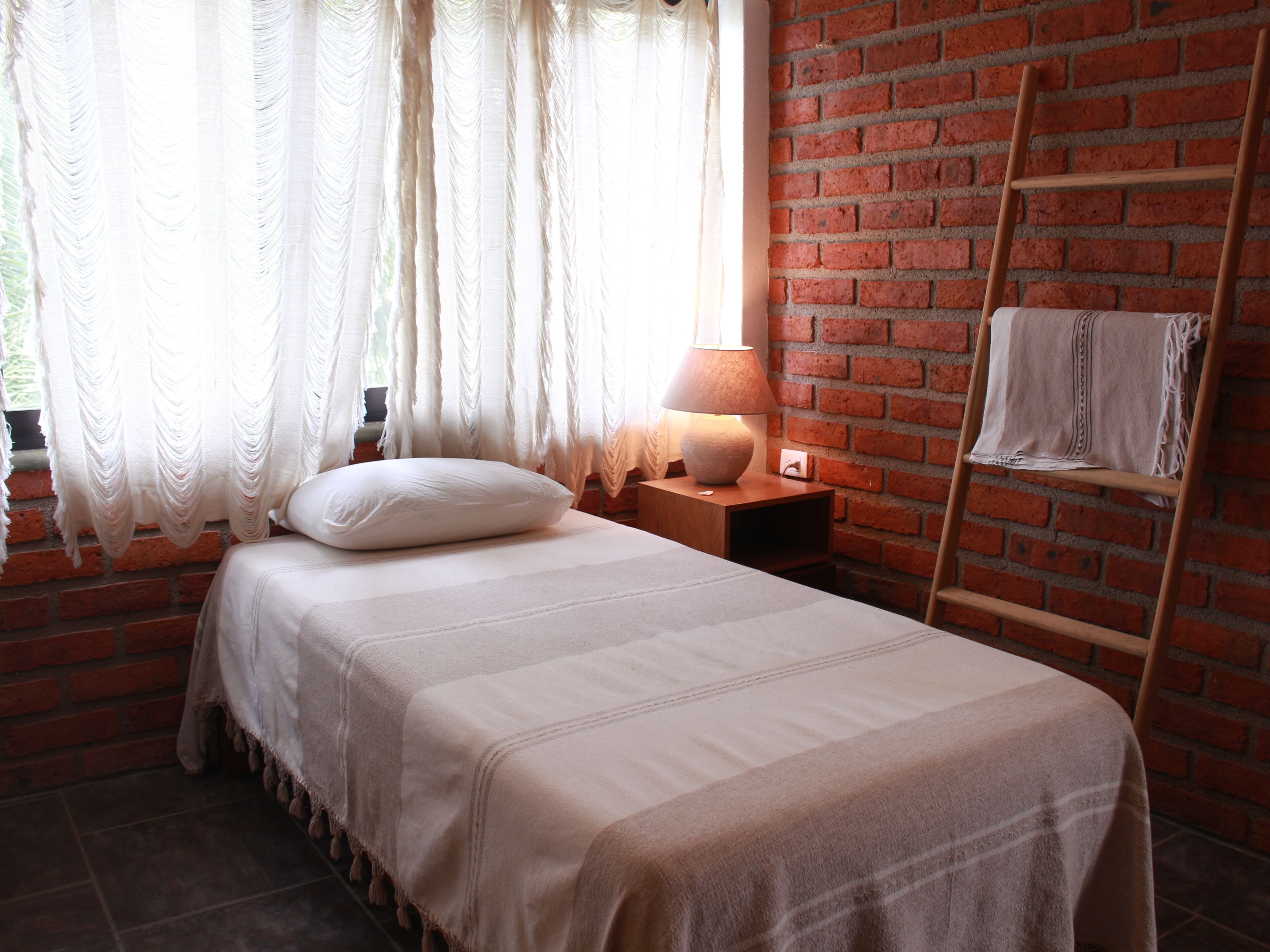 Un dormitorio con una cama, una mesita de noche, una lámpara, una ventana con cortinas y una pared de ladrillo.