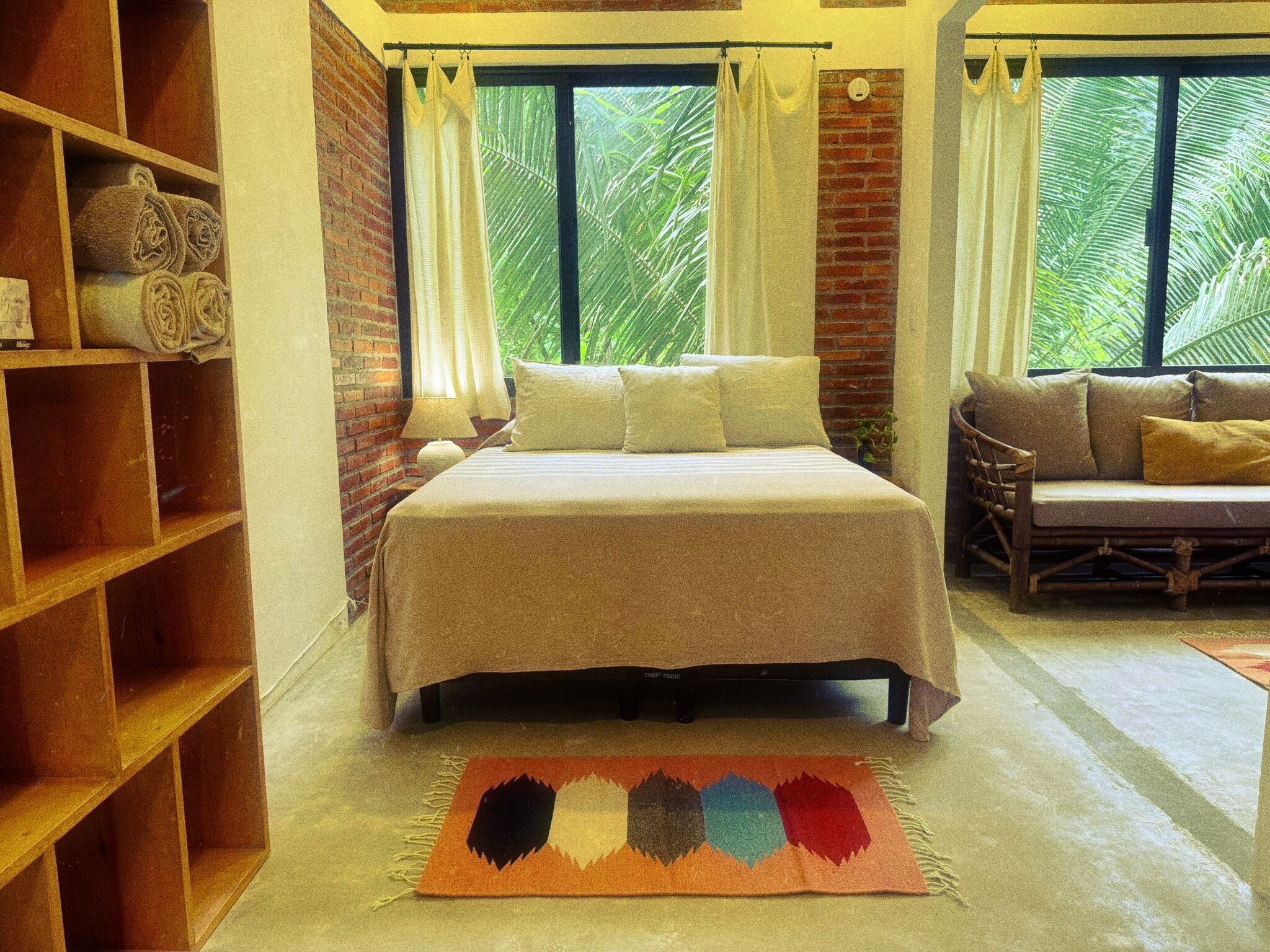A bedroom with a bed, a wooden shelf, a sofa, and a view of greenery outside.