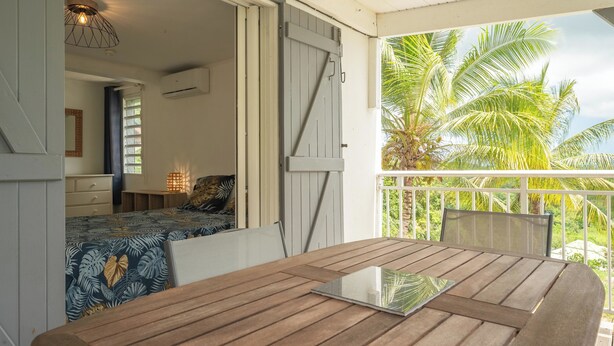 Une table en bois sur laquelle se trouve un ordinateur portable, un lit recouvert d’une couvre-lit à motifs, une fenêtre avec des rideaux et une vue sur des palmiers à l’extérieur.
