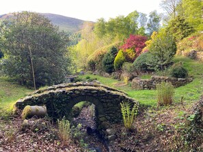 A stone arch bridge in a lush garden with diverse vegetation and a stone wall. - High Cross (Keswick)