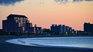 A coastal cityscape at dusk with a prominent hotel featuring multiple floors and balconies, a beach in the foreground, and a mix of residential and commercial buildings in the background. - Dazzling Oceanview-4 Train Stop Logan Airport (Revere)