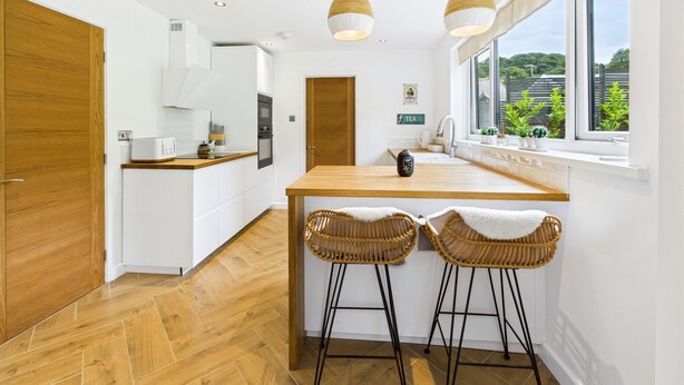 Une cuisine moderne avec un îlot central en bois, deux tabourets de bar en osier et des luminaires suspendus.