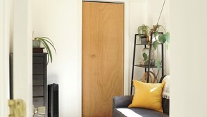 A room with a ceiling fan, a wooden door, a gray sofa with a yellow pillow, a black speaker, and a plant on a shelf. - Charming 3BR 2BA Home – Walk to Downtown St. Albans, Near Lake Champlain (Saint Albans City)