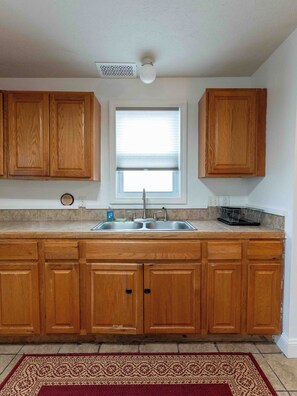 A kitchen with wooden cabinets, a double sink, a window with blinds, and a ceiling vent.