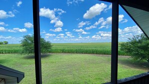 A view from a covered porch overlooking a cornfield and a grassy area with a clear blue sky and scattered clouds.