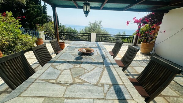 A stone-tiled patio with a central fire feature, surrounded by wooden chairs and potted plants.