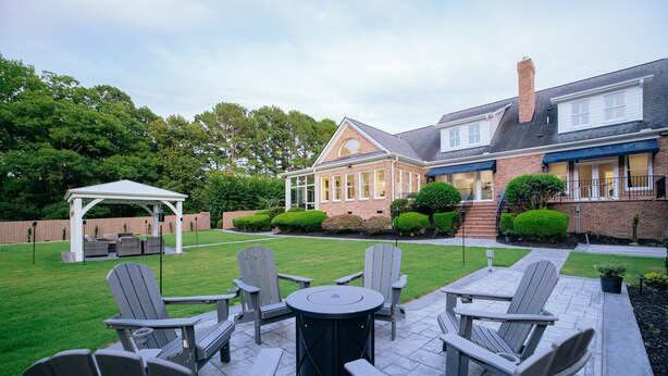 Une terrasse avec des fauteuils Adirondack et une table ronde, entourée d’une maison en briques dotée d’un espace couvert et d’une pelouse bien entretenue.