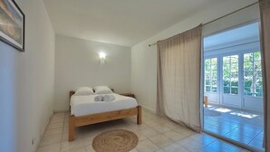 Une chambre avec un lit, une table de chevet, une lampe fixée au mur, une grande porte-fenêtre coulissante et une vue sur la verdure à l’extérieur.