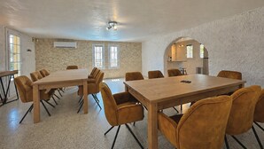 Een ruime kamer met een grote houten tafel en bijpassende stoelen, een stenen muur en een keukenruimte zichtbaar door een rondboogvormige doorgang.