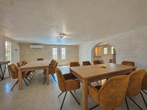 Une pièce spacieuse avec une grande table en bois et des chaises assorties, un mur en pierre et un coin cuisine visible à travers une porte en arc de cercle.