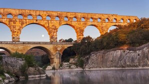 Un pont-aqueduc en pierre enjambe une rivière paisible dont les eaux claires se reflètent, le tout étant entouré d’un paysage rocheux et d’arbres.