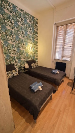 Une chambre avec deux lits, un plancher en bois, une fenêtre avec des stores et un mur orné d’un motif végétal.