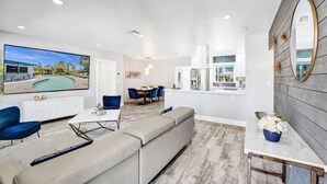 Una sala de estar moderna con un televisor de pantalla plana grande, un sofá, un comedor con sillas azules y una cocina con armarios blancos.