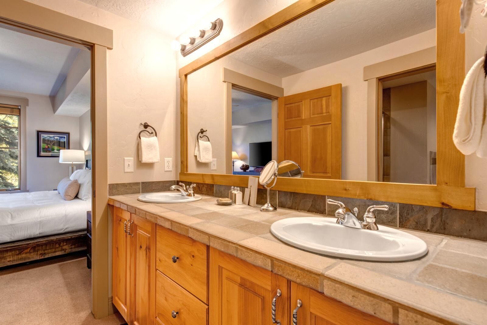 A bathroom with two sinks, wooden cabinets, a large mirror, and a view of a bedroom with a bed and window.