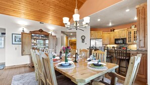 A dining area with a wooden table set for four, surrounded by wooden chairs. A chandelier hangs above the table, and a kitchen with wooden cabinets is visible in the background. - Town Pointe Collection by KBM - 2 Units (102c/301c) (Park City)