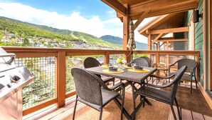 A covered outdoor dining area with wicker chairs and a grill, overlooking a scenic village and hills. - Town Pointe Collection by KBM - 2 Units (102c/301c) (Park City)