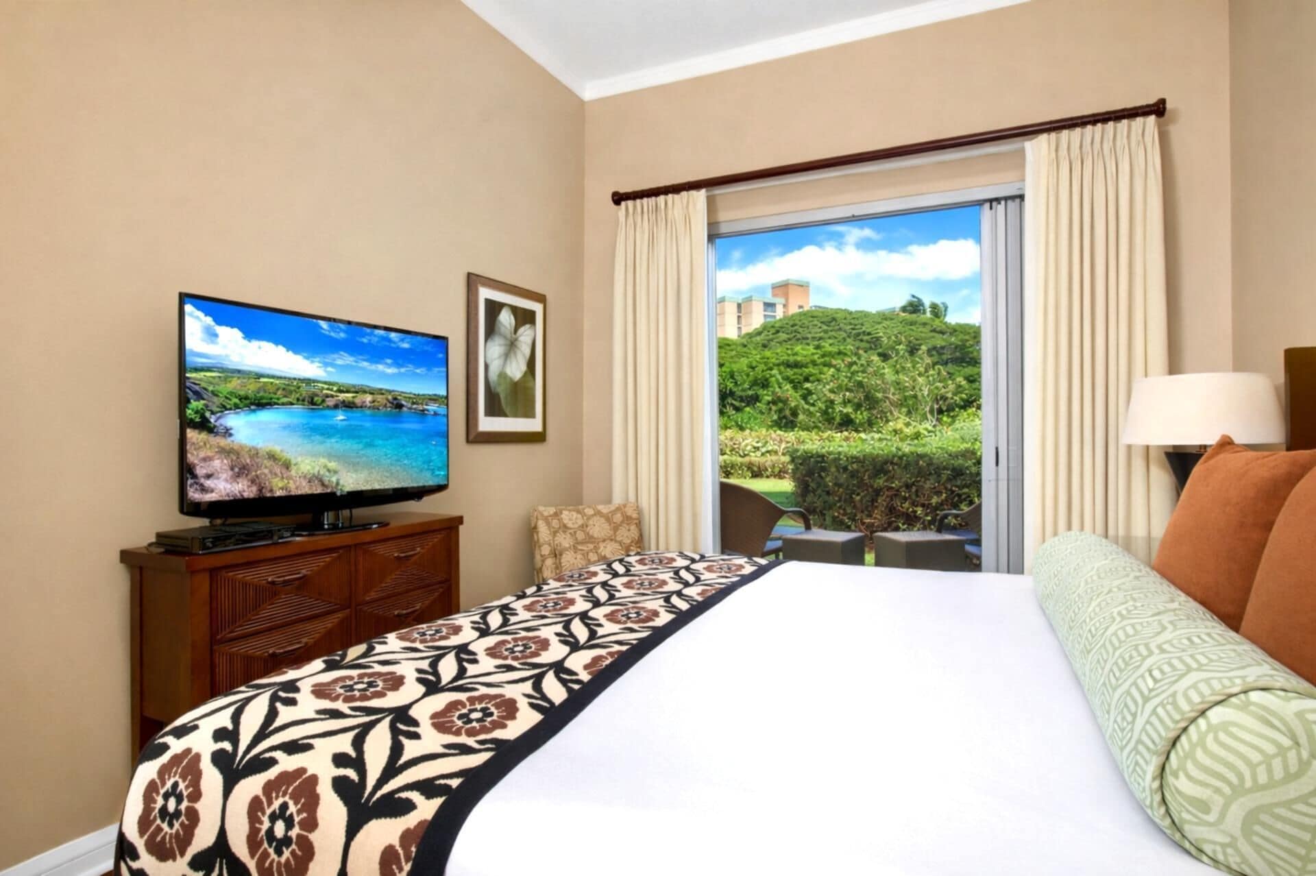 A hotel room with a bed, a television, a window with curtains, a view of greenery, and a lamp.