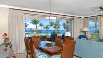 A dining area with a table and chairs, a sofa, a lamp, and a view of the ocean with palm trees.