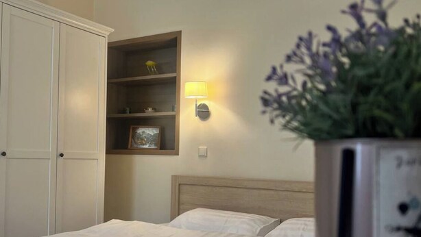 Une chambre d’hôtel avec un lit, une table de chevet avec une horloge, une étagère fixée au mur avec une lampe et une plante en pot.