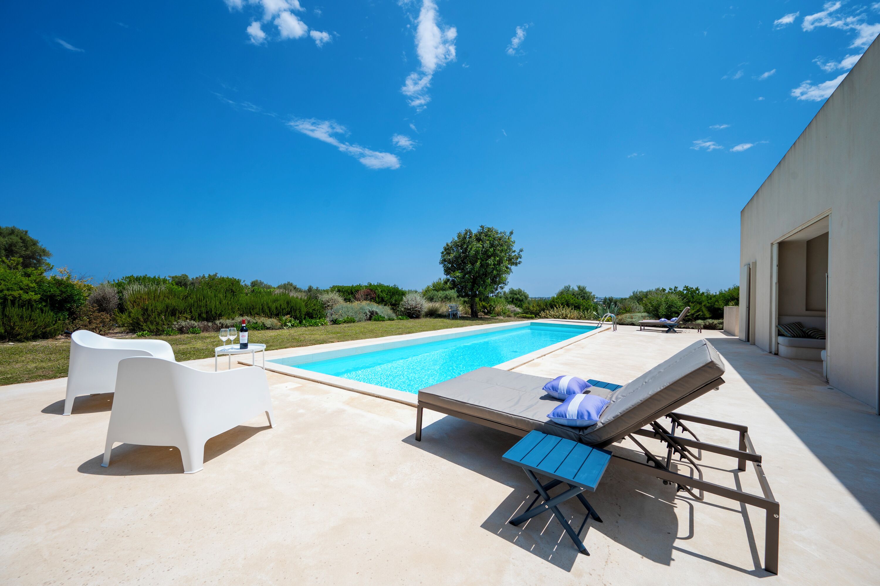 A swimming pool with lounge chairs and a table, surrounded by greenery and a clear sky.