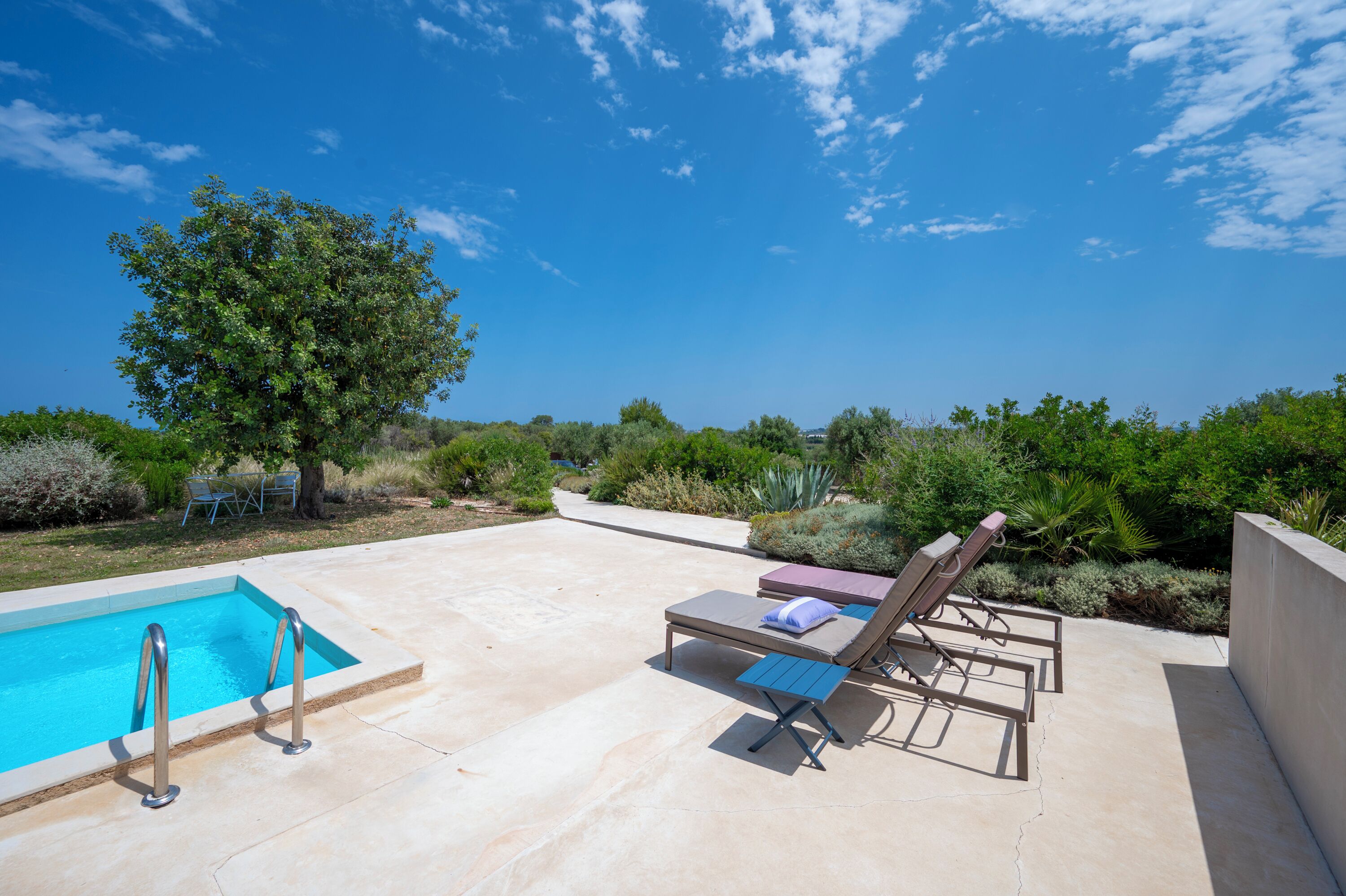 A pool area with two lounge chairs and a small table, surrounded by greenery and a clear sky.