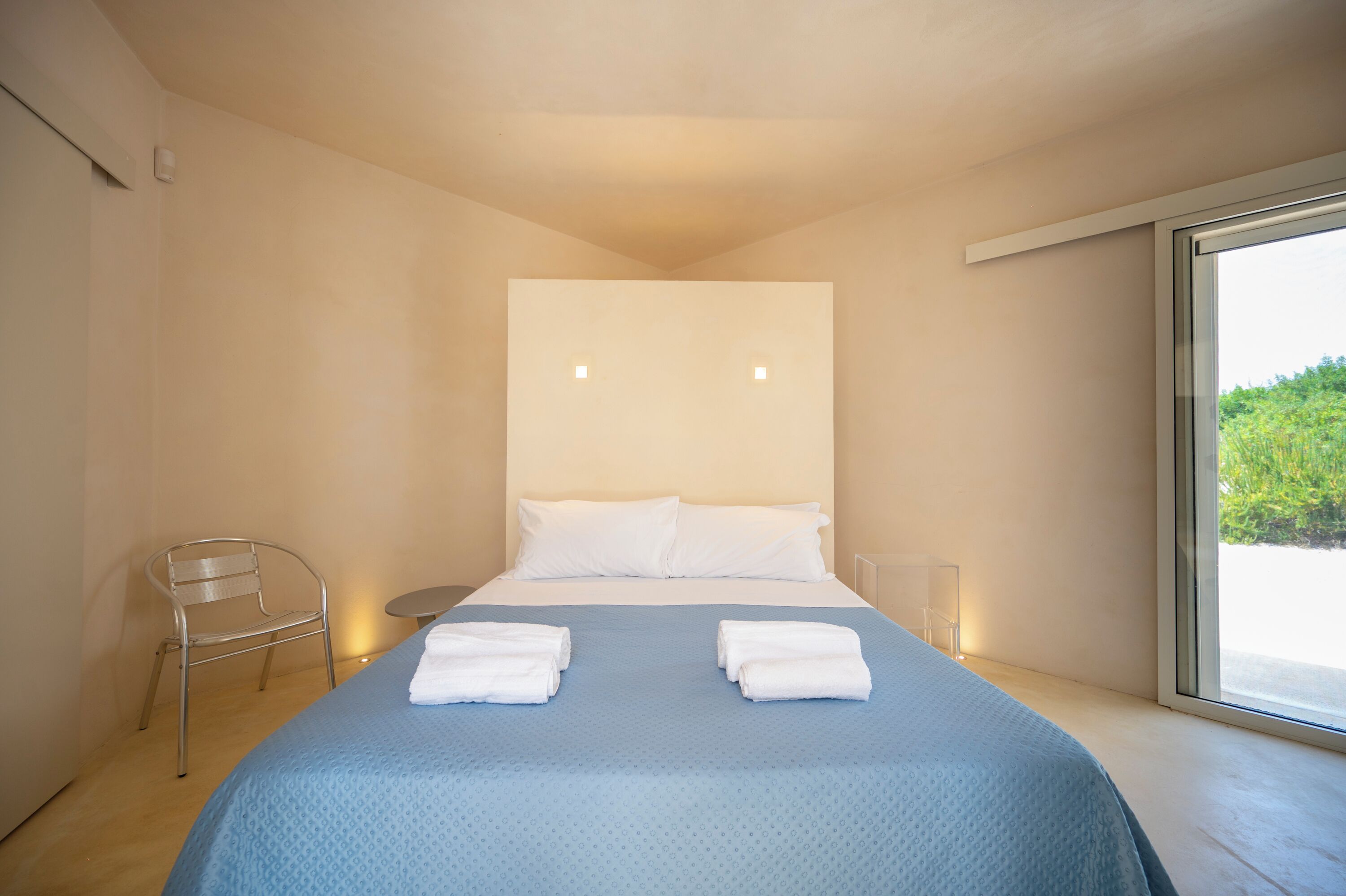 A modern hotel room with a large bed, two white pillows, a blue bedspread, a metal chair, and a large window offering a view of greenery.