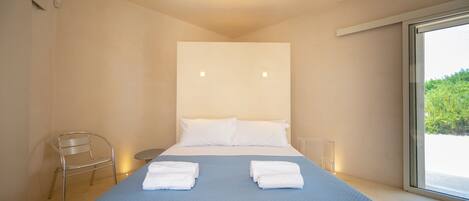 A modern hotel room with a large bed, two white pillows, a blue bedspread, a metal chair, and a large window offering a view of greenery.