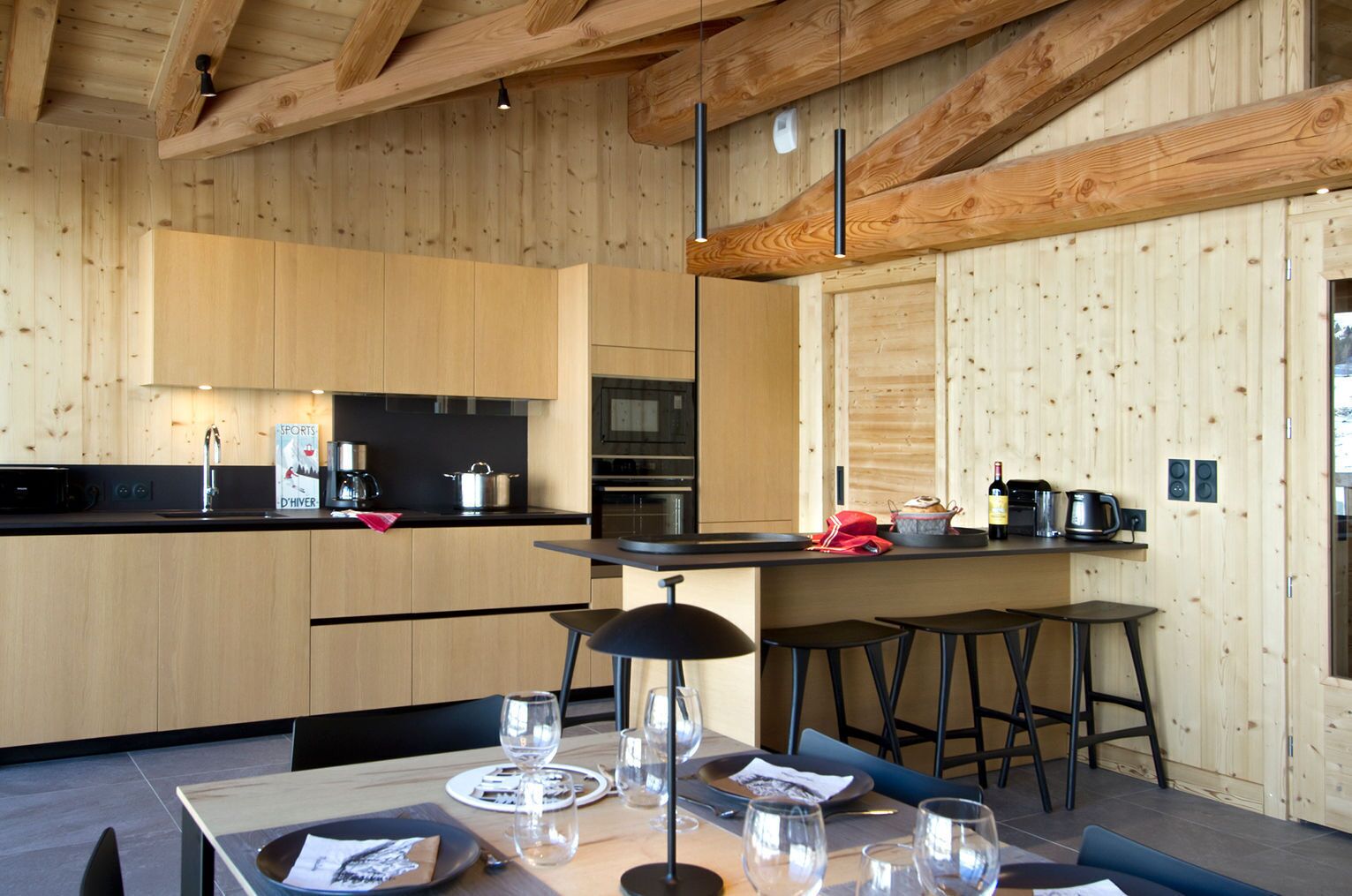 A modern kitchen with wooden cabinetry, a central island, bar stools, and a dining table set for two.