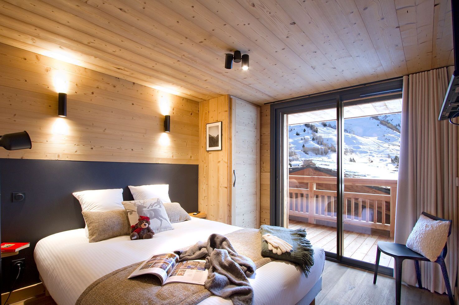 A cozy wooden room with a large bed, a nightstand, a reading lamp, and a view of a snowy landscape through a sliding glass door.