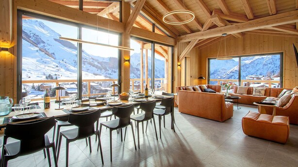 Une pièce spacieuse avec un plafond en bois, de grandes fenêtres offrant une vue sur les montagnes, un coin repas avec des chaises et des tables, et un espace détente avec des canapés et une table basse.