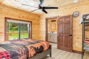 Een slaapkamer in een houten hutje, met een bed, een plafondventilator, een raam, een deur en een kleine kitchenette.