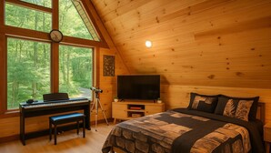 Ein gemütliches Schlafzimmer mit einem Bett, einem Schreibtisch mit Computer, einem Fernseher und einem Fenster mit Blick auf den Wald.