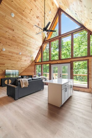A spacious loft with a high ceiling, wooden beams, a large window, a sofa, a kitchen island, and a TV.