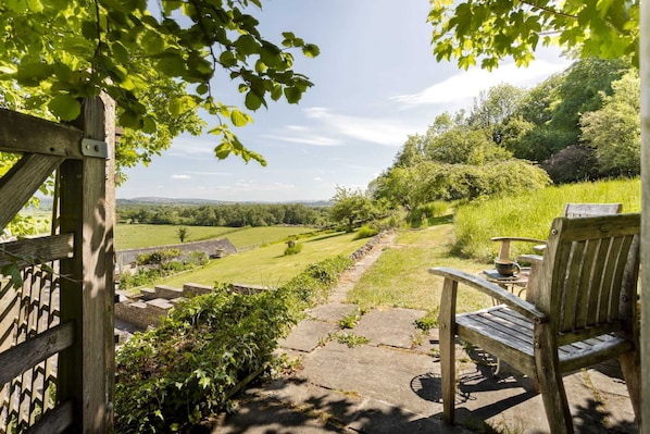 Un banc et une table en bois sont installés sur un chemin de pierre dans un jardin, offrant une vue sur un champ verdoyant et des arbres.