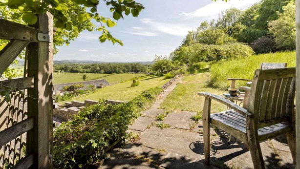 A wooden bench and table set on a stone pathway in a garden with a view of a green field and trees. - The Room Rawsons in Levens (Kendal)