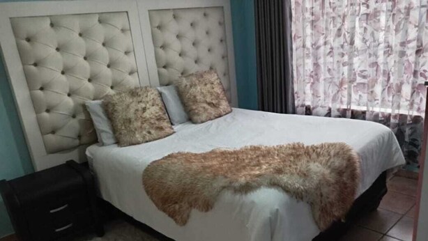 A bedroom with a tufted headboard, a bed with a white bedspread and a brown fur throw, a black nightstand, and a window with floral curtains. - Secure, Spacious & Fully Equipped – Your Ideal Townhouse Stay 4 (Pretoria)
