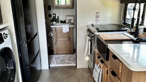 A compact kitchen with a washing machine, a refrigerator, a microwave, and a stove. There is a rug on the floor and a window providing natural light. - Modern Tiny Home • Firepit • Near AT&T Stadium (Venus)