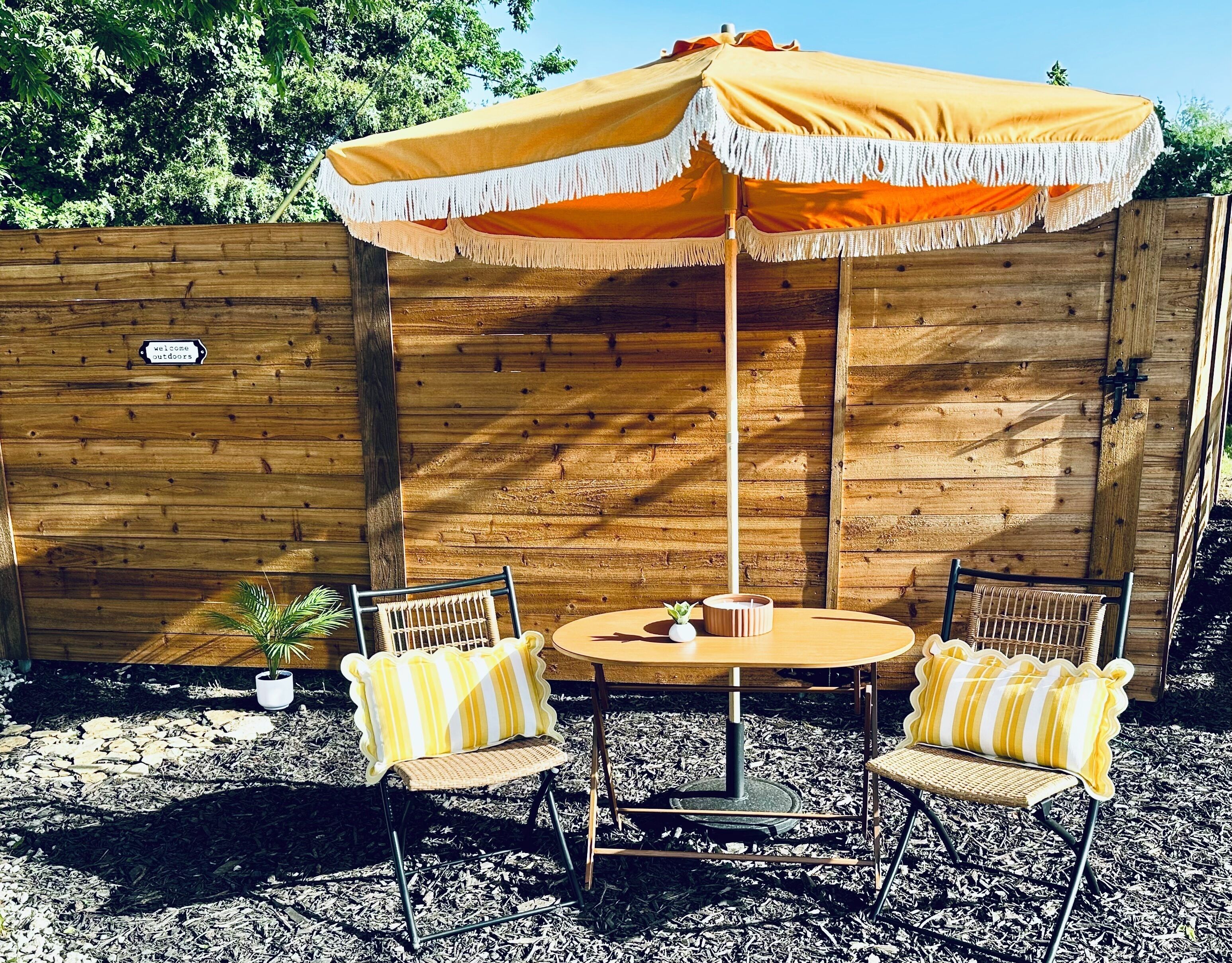 An outdoor seating area with a wooden fence, two wicker chairs with yellow and white striped cushions, a round table, and a yellow and white striped umbrella.
